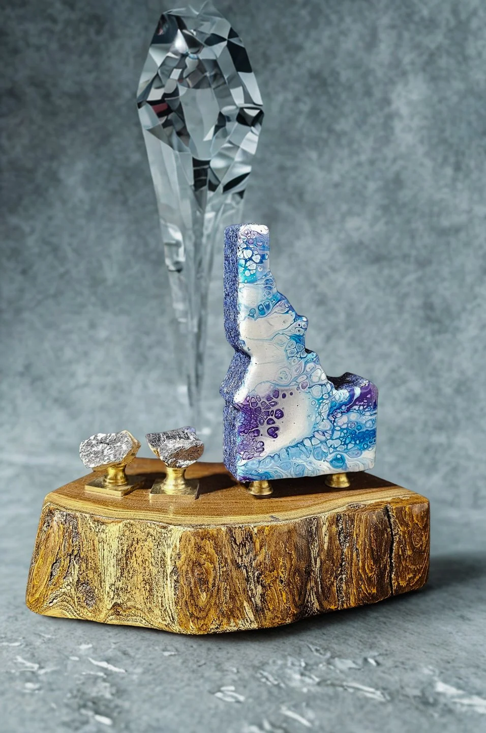 Decorative art piece featuring a large crystal, a colorful abstract sculpture, two small metallic rocks on brass supports, all on a textured wooden base.