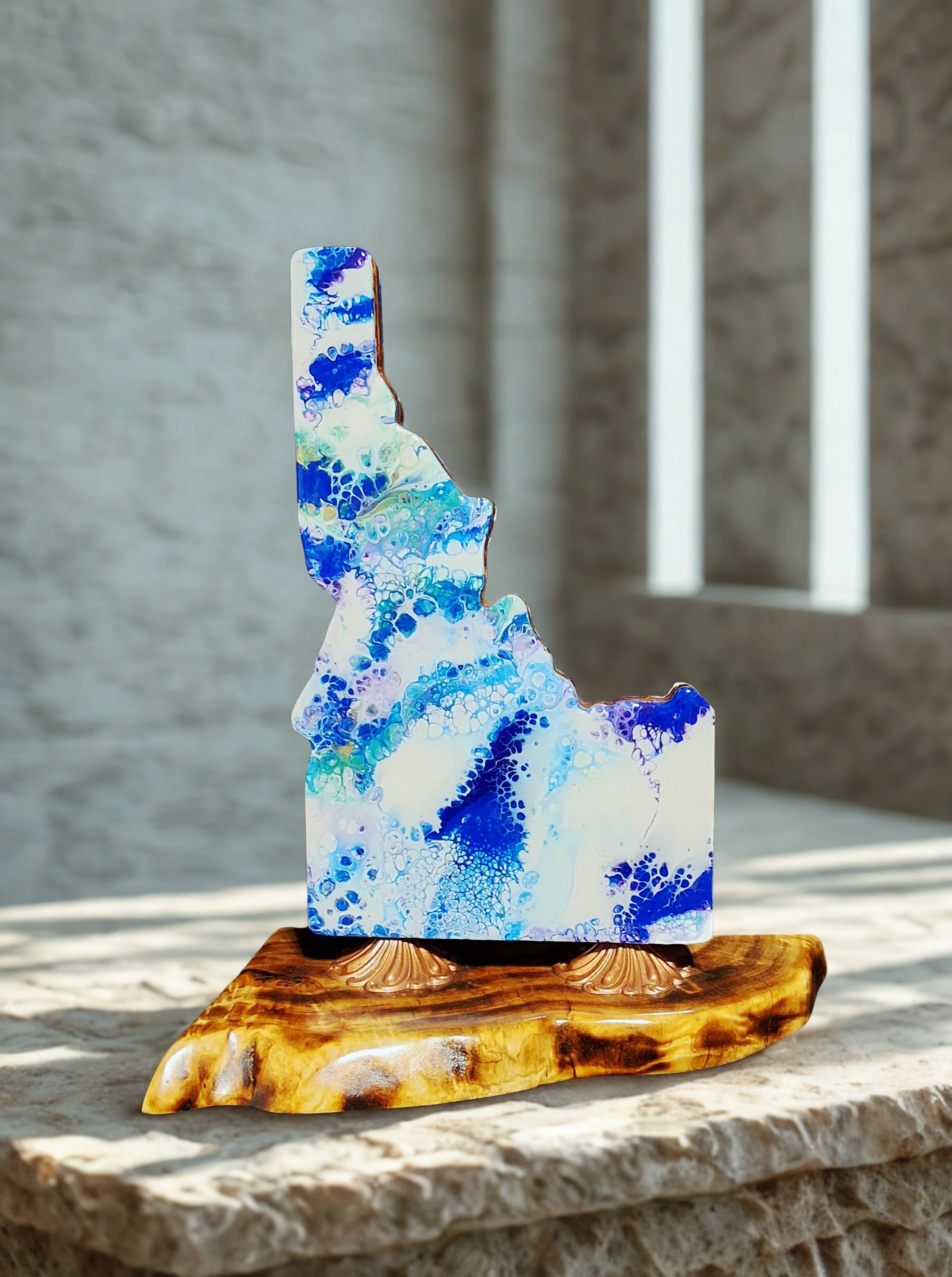 Glass sculpture with blue and white marbled patterns, mounted on a polished wooden base, set on a stone surface with natural light and shadows.