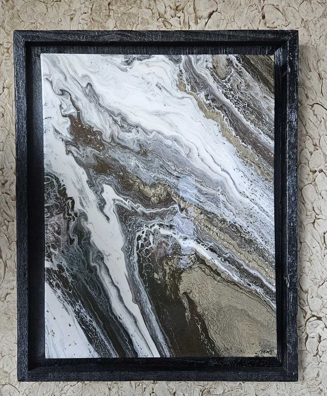 Abstract marbled painting with black, white, gold, and gray swirling patterns inside a black frame, placed on a textured beige surface.