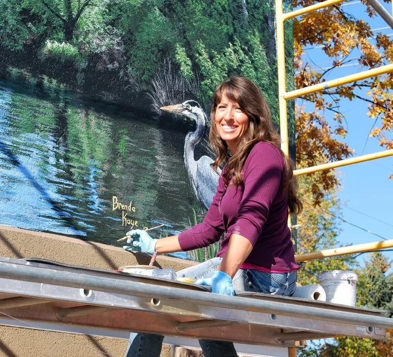 Woman painting a mural of a heron by a river, wearing a purple long sleeve shirt and blue gloves, outdoors on a sunny day.