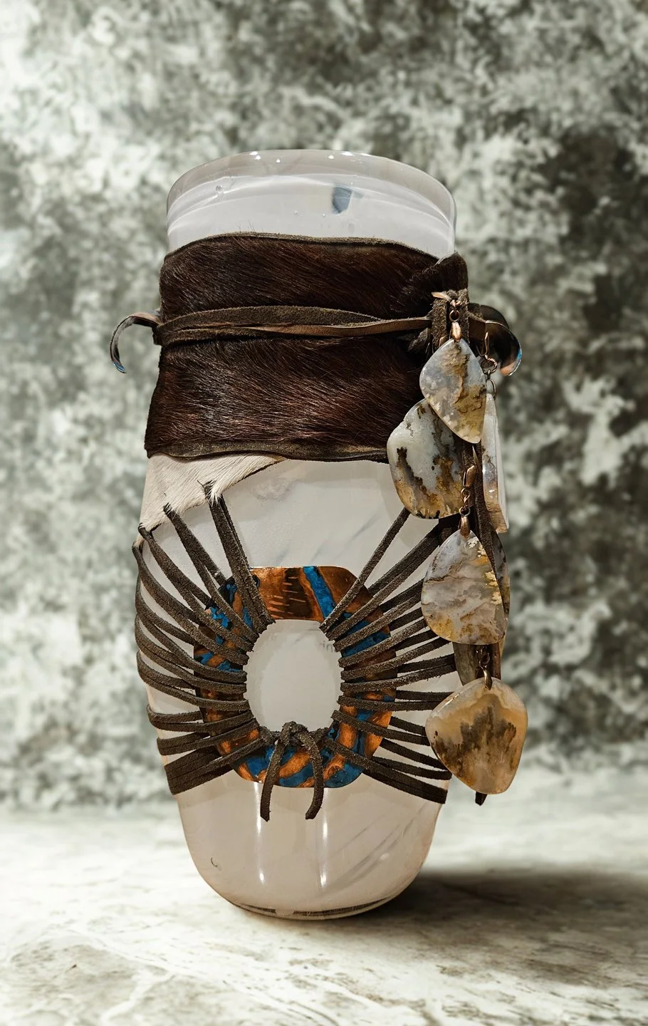 A clear glass jar decorated with leather straps, shells, and a piece of animal hide, with a dark brown leather strap wrapped around the top and some shell charms hanging on the side, set against a blurred gray background.