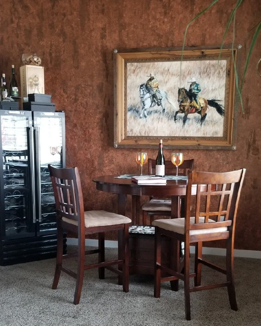 A small round wooden dining table with three chairs, two with beige cushions, two glasses of white wine, a bottle of wine, and some papers on the table. A painting of two cowboys riding horses hangs on a textured brown wall behind the table. To the l