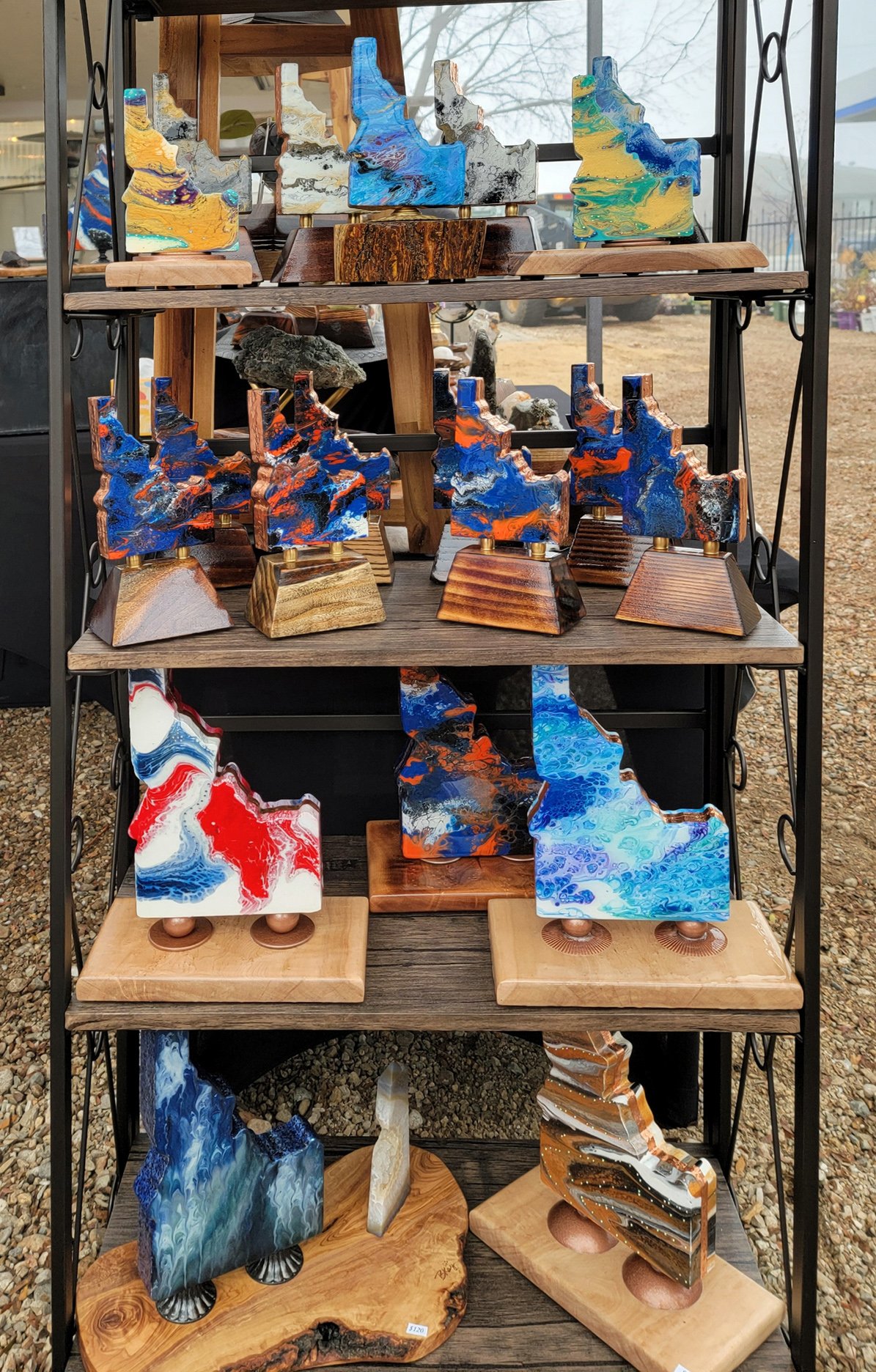 Colorful abstract sculptures displayed on a black metal shelf with wooden base in a craft fair setting.
