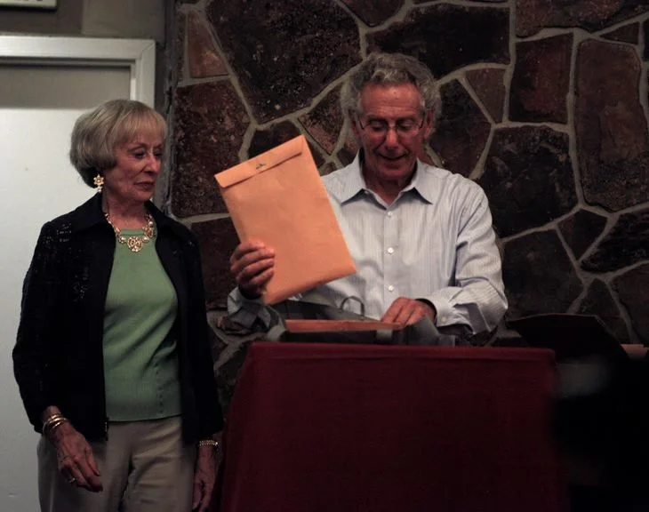 A woman and a man are standing behind a table indoors, with the man holding a large manila envelope and looking down at something on the table. The woman is observing, wearing a black blazer and a green top with jewelry, while the man is dressed in a