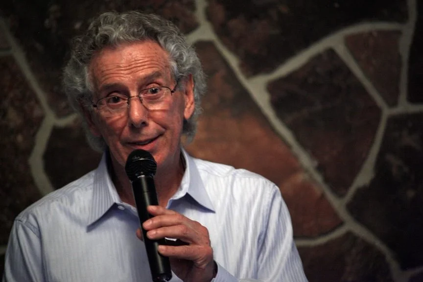 An elderly man with gray, curly hair and glasses, wearing a light blue collared shirt, speaking into a microphone against a brick and stone wall background.