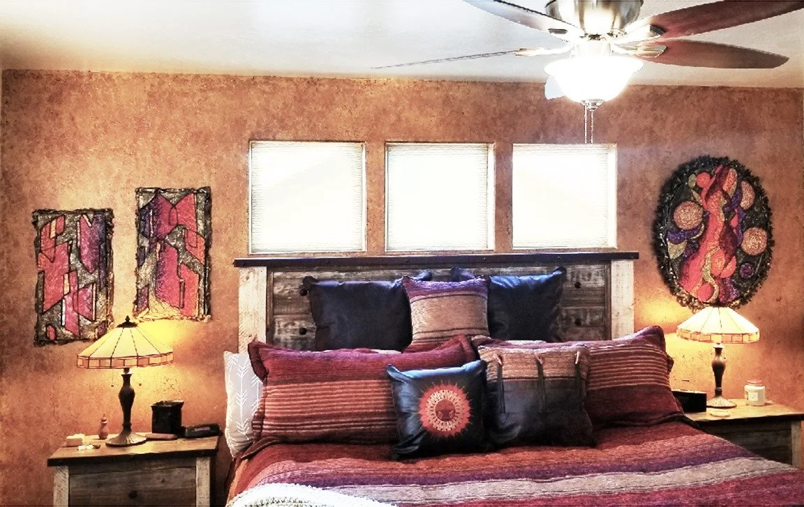 A bedroom with a rustic wooden bed, multiple pillows, and matching nightstands with lamps. The wall behind has two small windows, framed artwork, and a decorative piece.