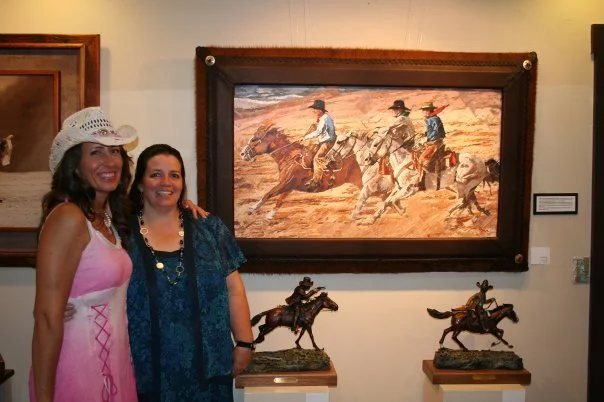 Two women standing in front of artwork and sculptures at an art gallery. One woman is wearing a white hat and pink dress, the other a blue dress. The artwork depicts cowboys riding horses in a desert setting.