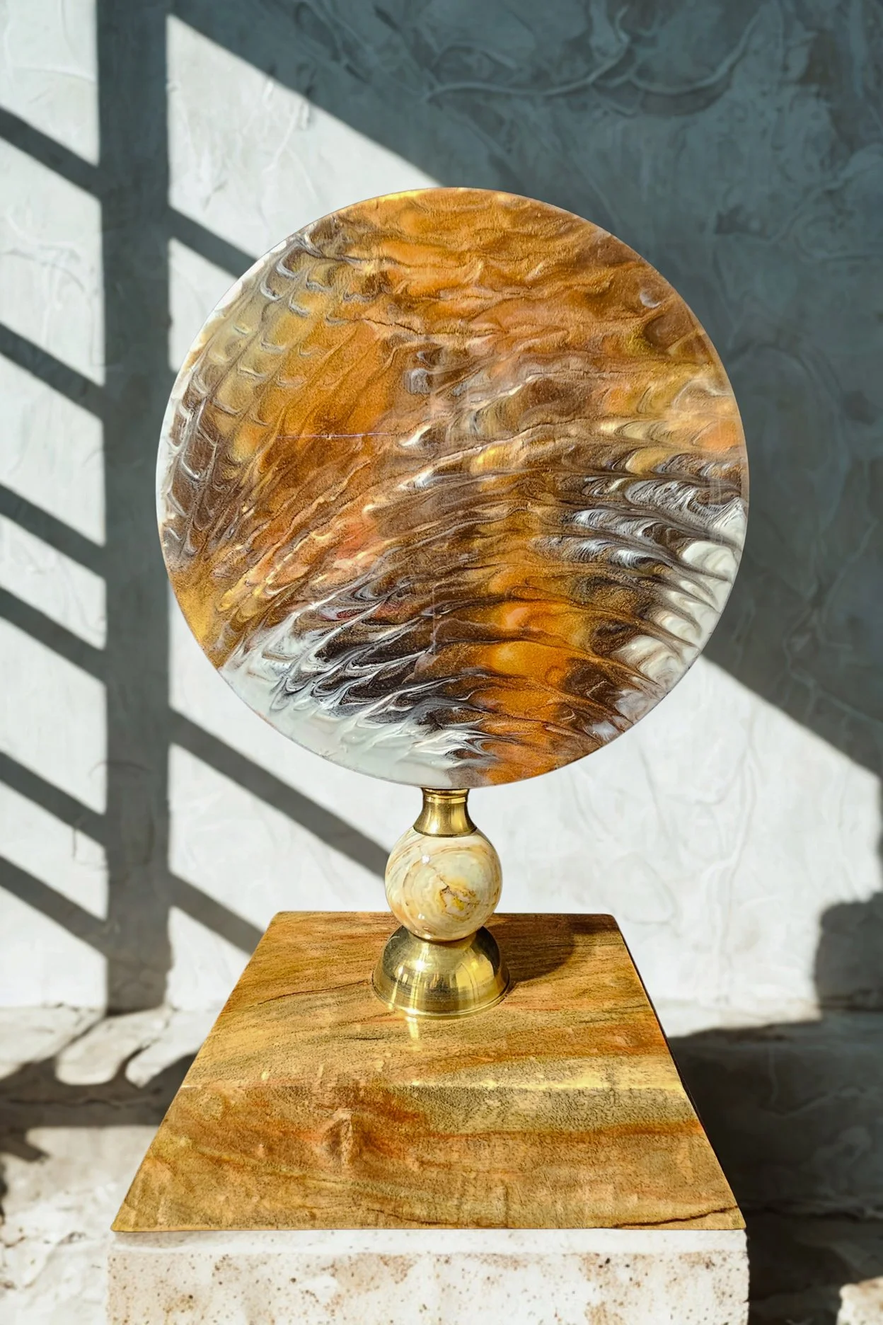 Decorative glass art sculpture with swirling amber, brown, and white colors, mounted on a marble and brass stand, placed on a wooden surface outdoors with sunlight and shadow patterns.