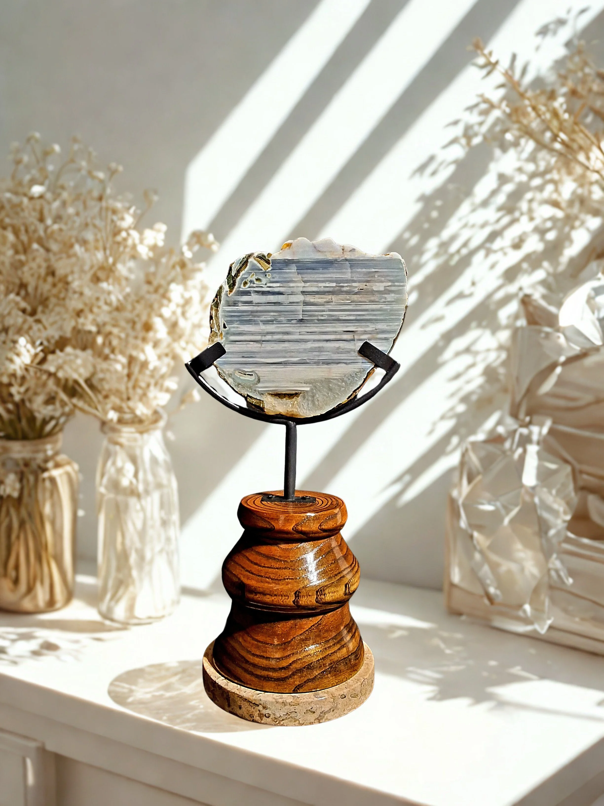 A decorative piece featuring a round polished geode or mineral on a black metal stand, mounted on a carved wooden base, placed on a white surface with dried flowers in the background and sunlight casting shadows.