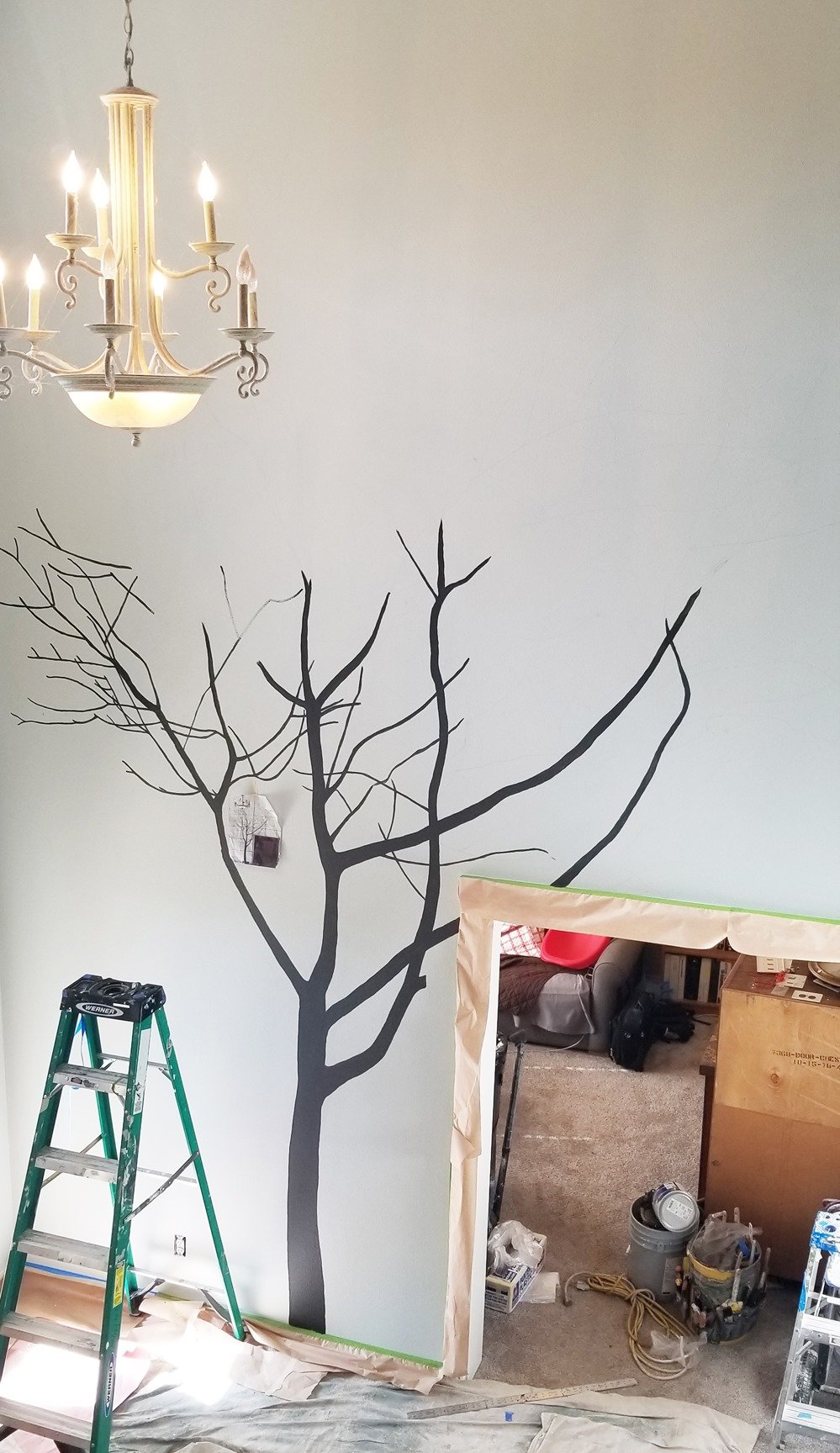 A partially painted wall with a black silhouette of a leafless tree and a chandelier hanging from the ceiling, with an opening in the wall leading to a room with construction supplies.