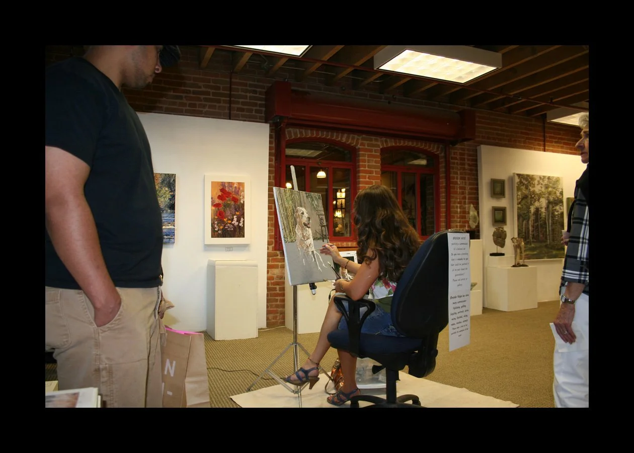 A woman is painting in an art gallery while two people observe. The gallery has brick walls, artwork displayed, and a woman sitting on a chair with a painting easel in front of her.