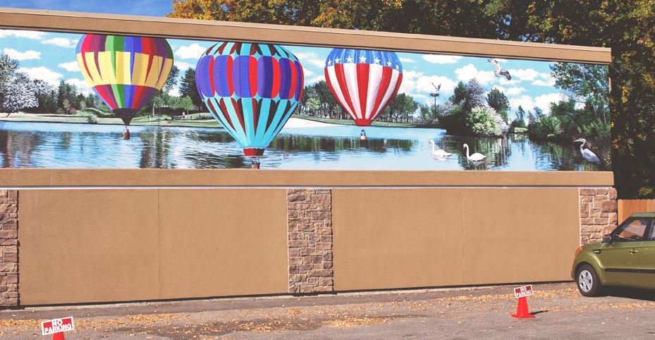 Large billboard showing a digital painting of hot air balloons floating over a lake with swans, trees, and a blue sky with clouds, mounted on a brick wall, with a car parked nearby.
