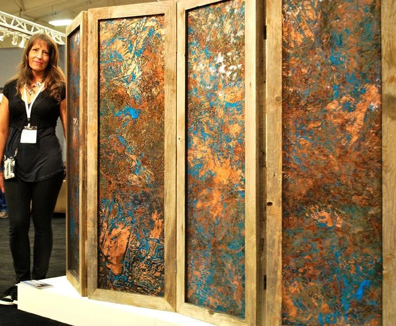 A woman standing beside a large, multi-pane abstract artwork with rust, blue, and gold colors on a wooden display.