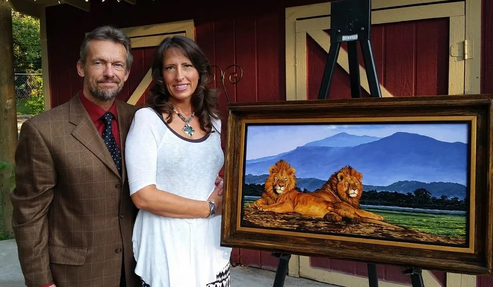 A man and woman standing together next to a framed painting of two lions lying on a log with mountains in the background.
