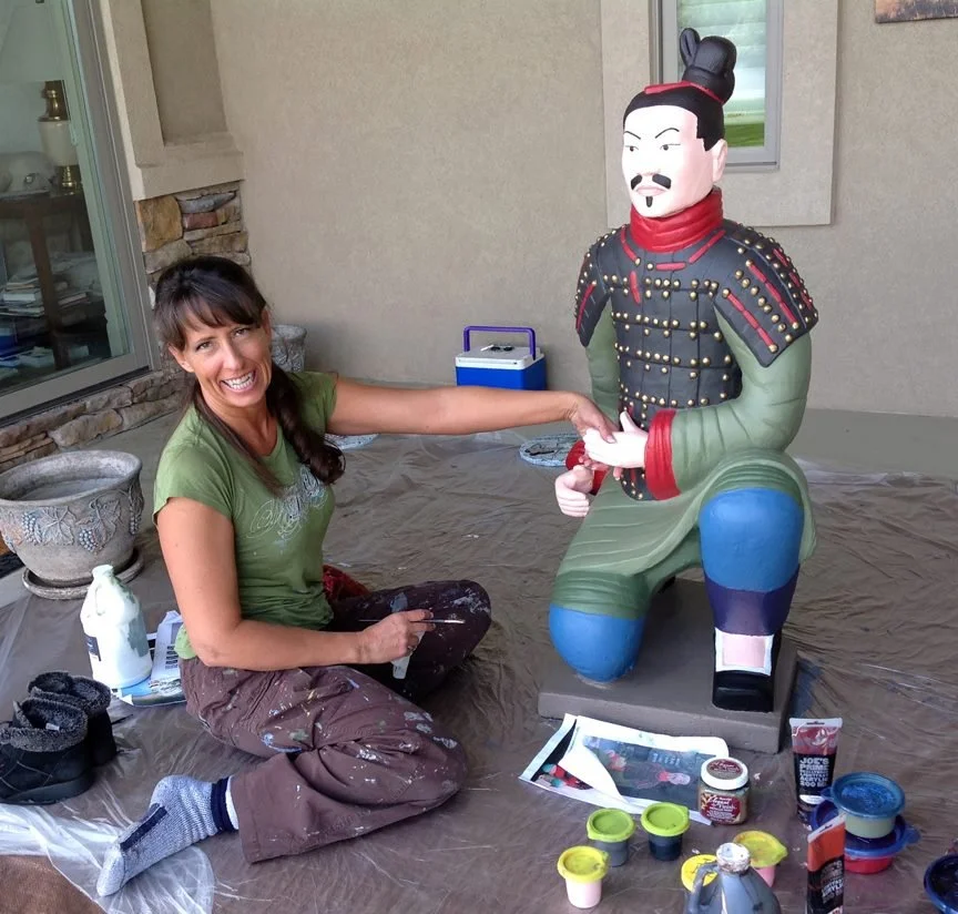 Woman painting a large colorful statue of a martial artist in traditional attire, kneeling on a protective sheet outdoors.