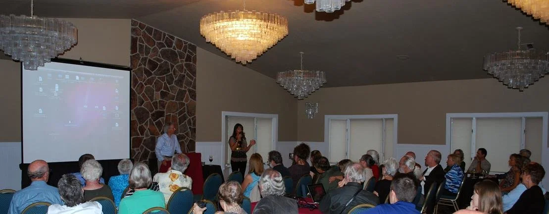 A woman speaking at a podium during a presentation in a banquet hall with chandeliers, with an audience seated facing her.