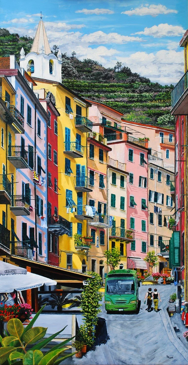 Colorful Mediterranean street scene with multicolored buildings, balconies, laundry hanging to dry, a small green bus, and two people walking. Hills and a church steeple are in the background under a partly cloudy sky.