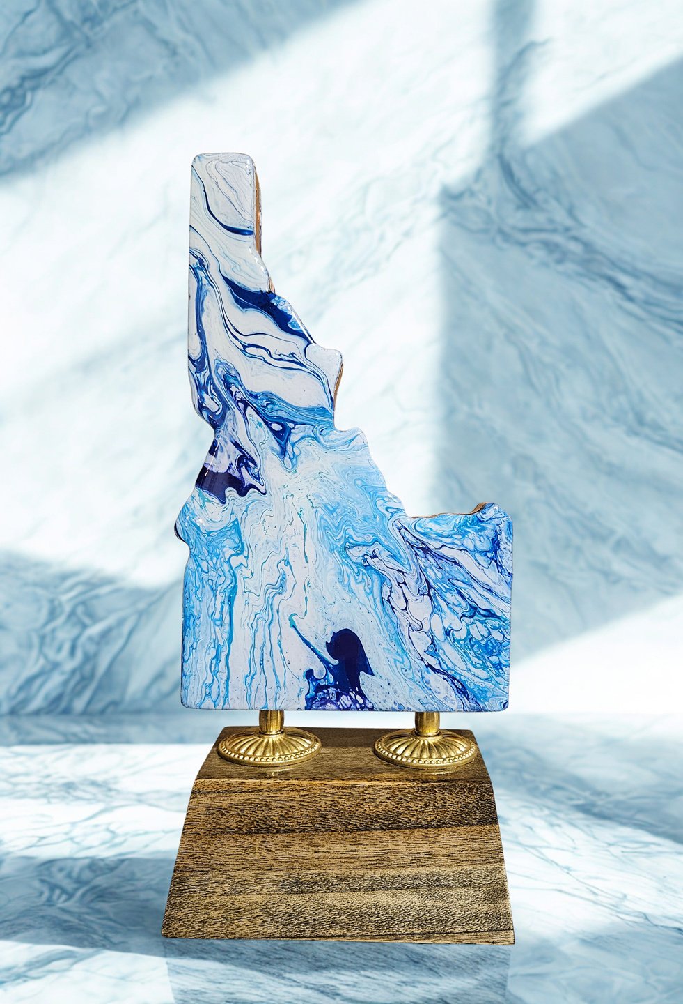 An abstract art piece with swirling blue, white, and black patterns mounted on two gold stands, placed on a wooden base, against a white background.