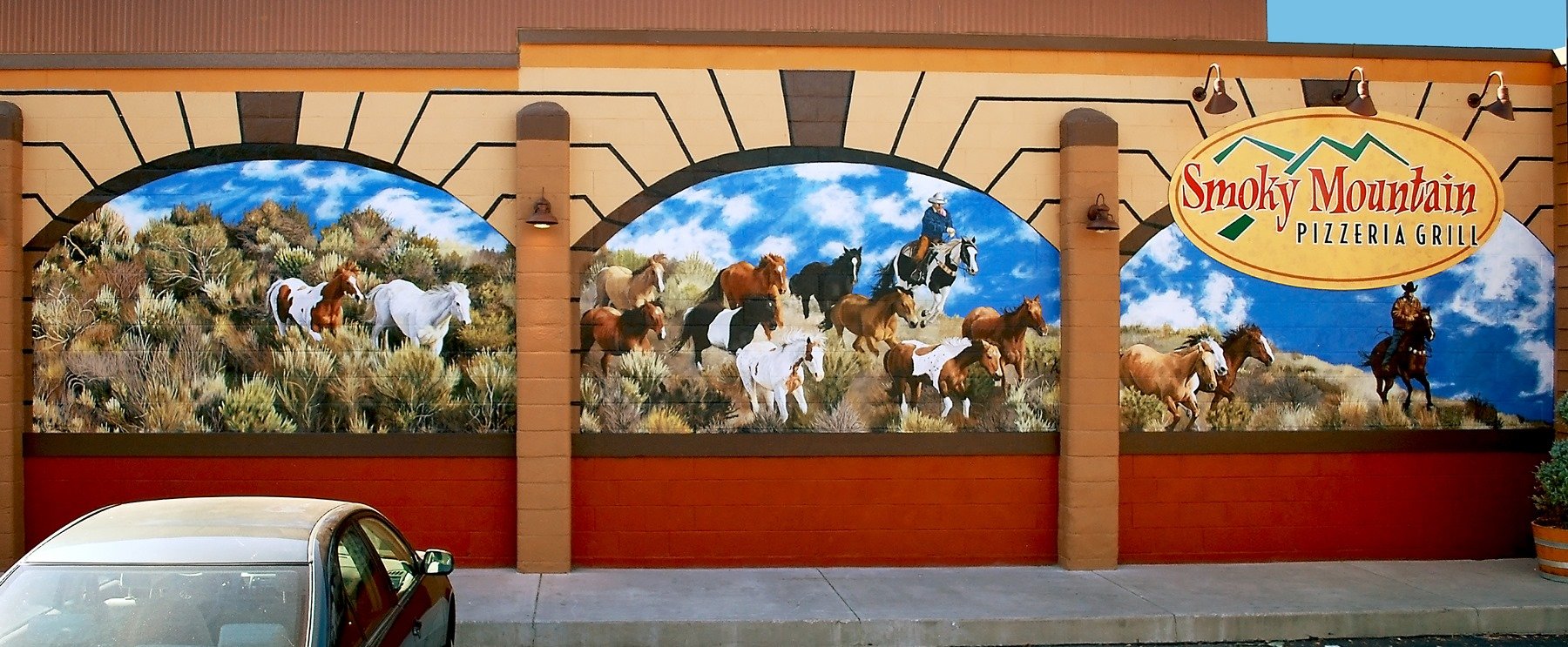Colorful mural of horses in a desert landscape at Smoky Mountain Pizzeria Grill, with a cowboy riding a horse and a sheriff on a horse, and the restaurant's sign in the top right corner.