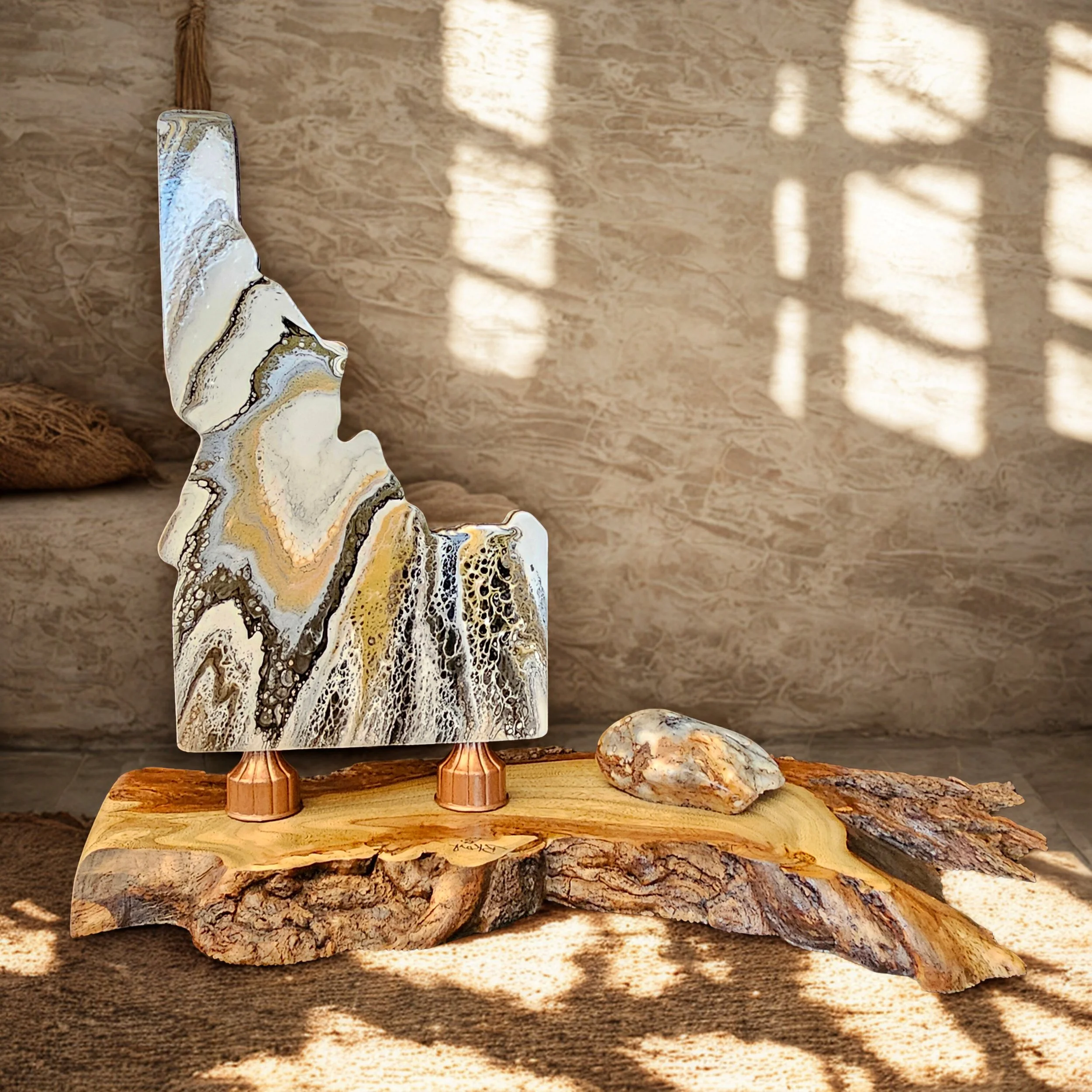 Decorative abstract sculpture with swirling black, white, and gold patterns, placed on a natural wooden base with a small rock beside it, illuminated by sunlight casting shadow patterns on the wall.