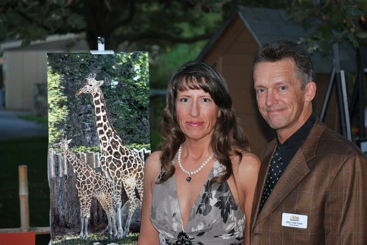 A man and woman standing outdoors next to a poster of giraffes, with trees and a small shed in the background.