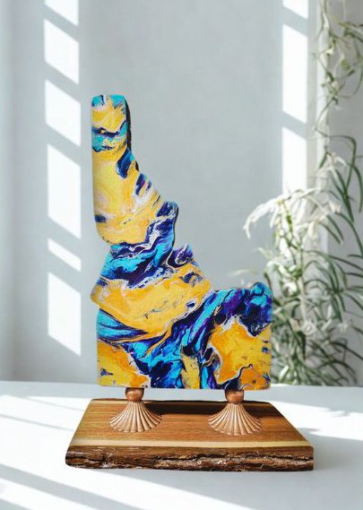Abstract acrylic painting in blue, yellow, and black on a resin sculpture, displayed on a wooden base with two small decorative feet, in a bright room with sunlight and plants.