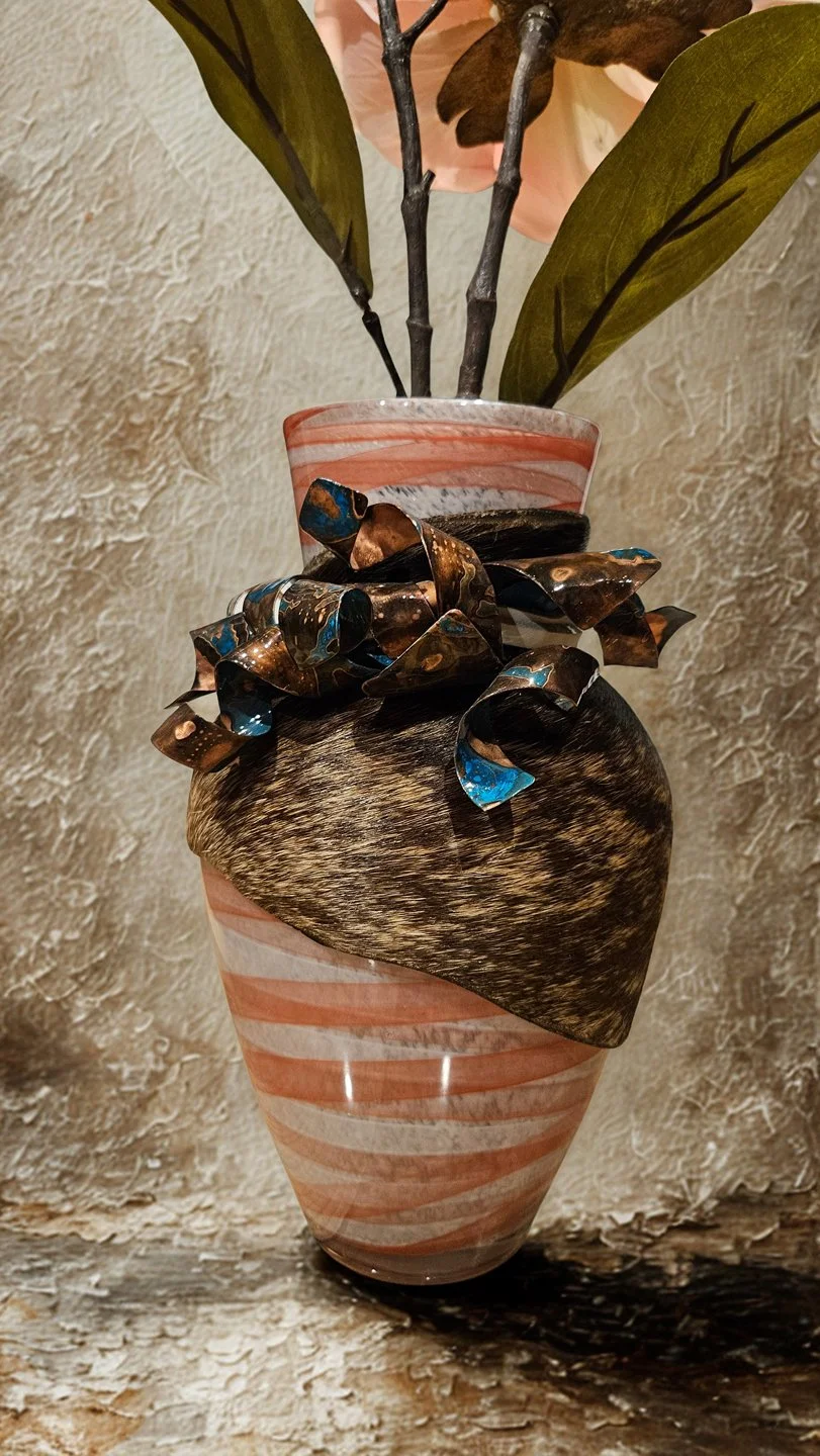 Decorative vase with green leaves, a large dark feather, and a feathered ornament, placed on a textured surface against a stone wall background.