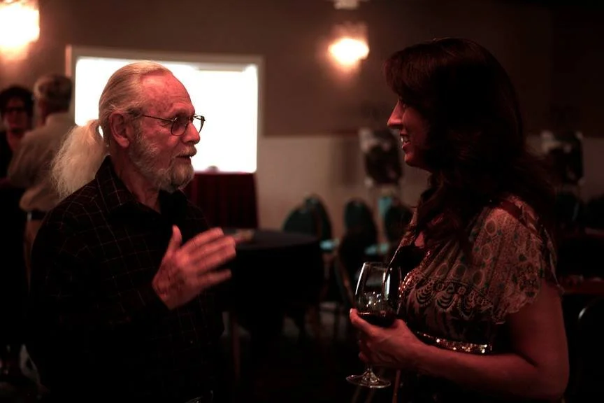 Two women engaged in a conversation at a dimly lit social gathering, one with long gray hair and glasses, the other holding a glass of red wine.