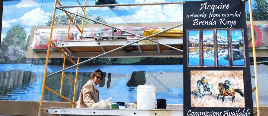Woman in tan jacket at art booth with landscape paintings and a sign offering mural artwork commissions.
