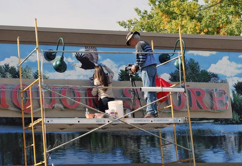 Two people working on a mural painting on a wall, with scaffolding and painting supplies, under a partly cloudy sky with trees in the background.