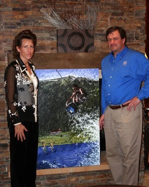 A woman and man standing beside a large landscape painting with water, mountains, and sky, in front of a brick wall decorated with a vase and dried branches.