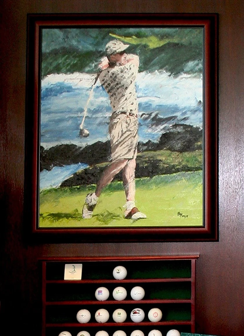 A framed watercolor painting of a man playing golf near water and rocks, with a set of golf balls arranged on a shelf below.