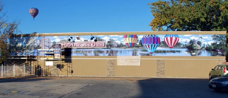 Large billboard mural depicting hot air balloons over a lake with trees and swans, with a parking lot in the foreground.