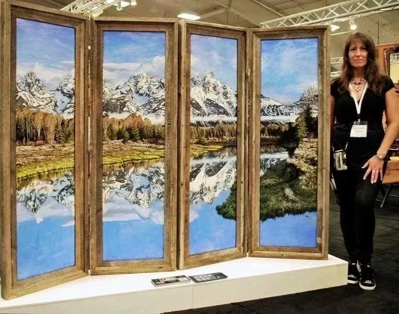 A woman standing next to a folding room divider with landscape paintings of snow-capped mountains, trees, and a lake reflecting the mountains.