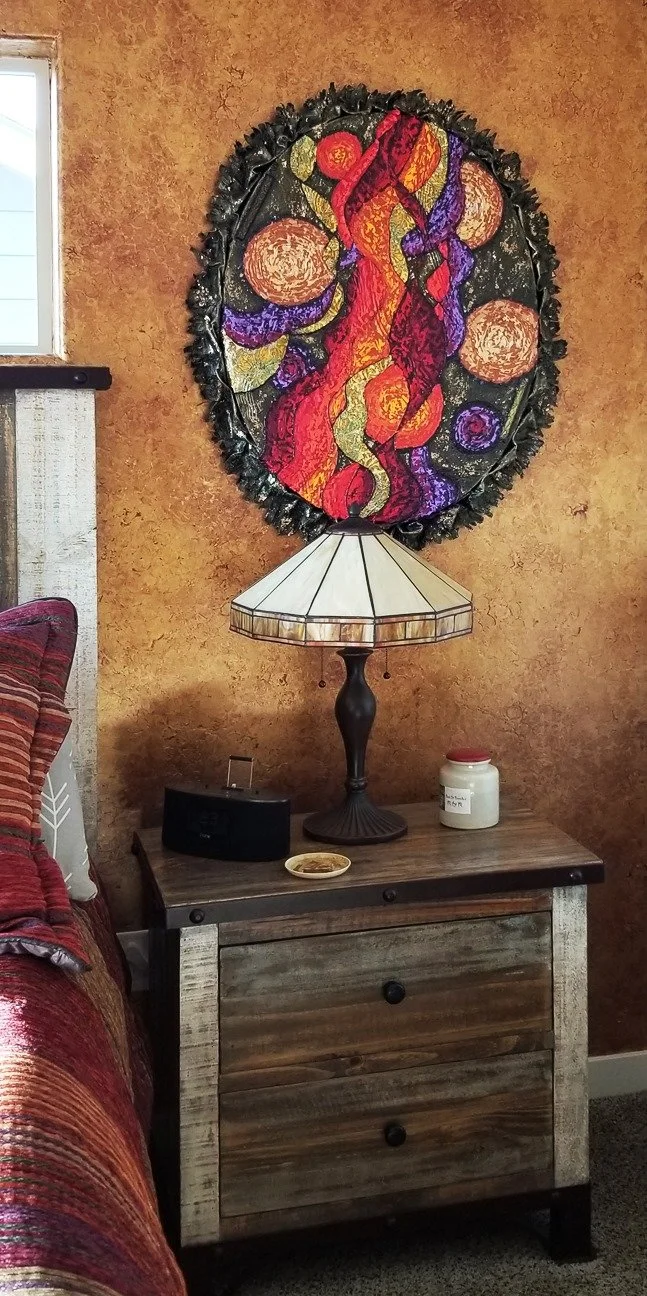 Decorative oval painting with abstract figures and circular patterns hanging above a wooden bedside table with a lamp, a small black speaker, a candle, and a decorative dish in a cozy room with rust-colored walls.