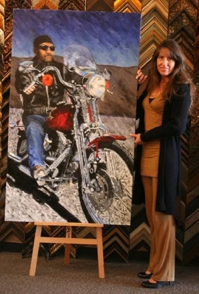 A woman standing next to a large painting of a man on a motorcycle. The man in the painting is wearing sunglasses, a black jacket, and jeans, with a desert landscape in the background.