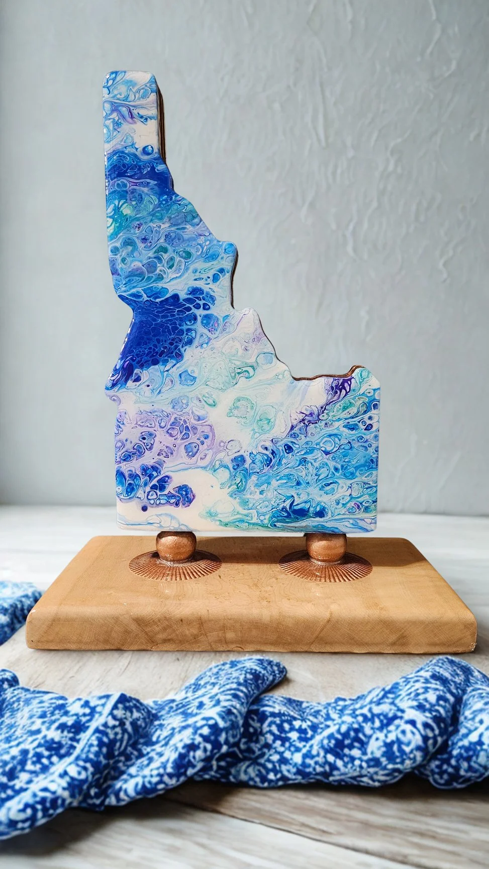 A decorative abstract art piece with blue, purple, and white swirls on a shape resembling a state or a landmass, mounted on a small wooden base with two copper-colored supports.