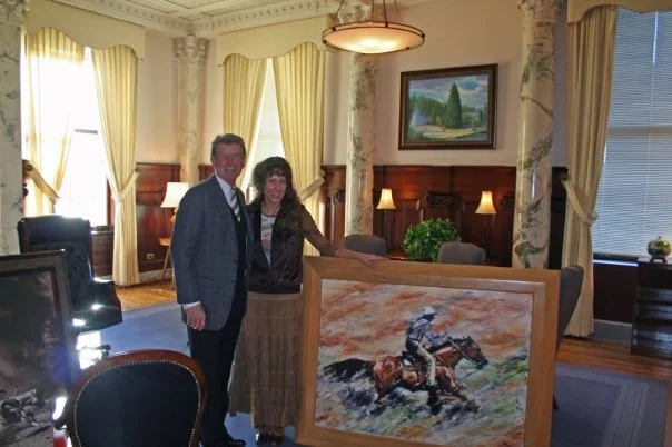 A man and woman standing next to a framed painting of a horse and rider in a well-lit room with large windows and elegant curtains.