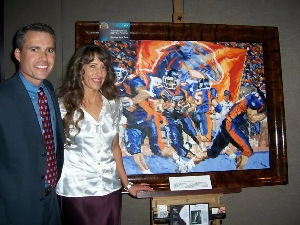 A man and woman stand next to each other beside a framed painting of a football game with players in blue and orange uniforms.