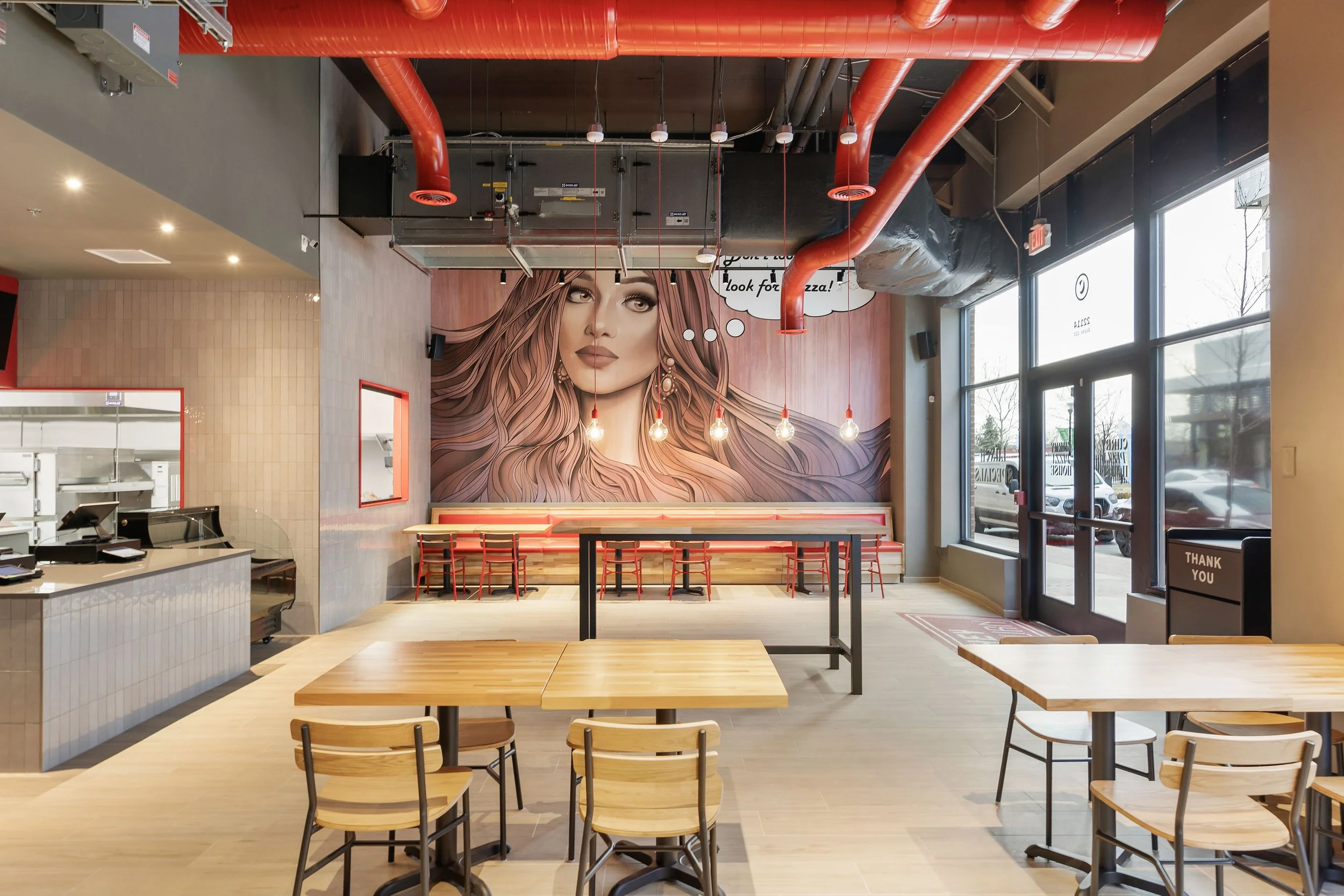 Inside a modern pizza restaurant with wooden tables and chairs, a large mural of a woman with long hair and makeup on the back wall, and red ductwork on the ceiling.