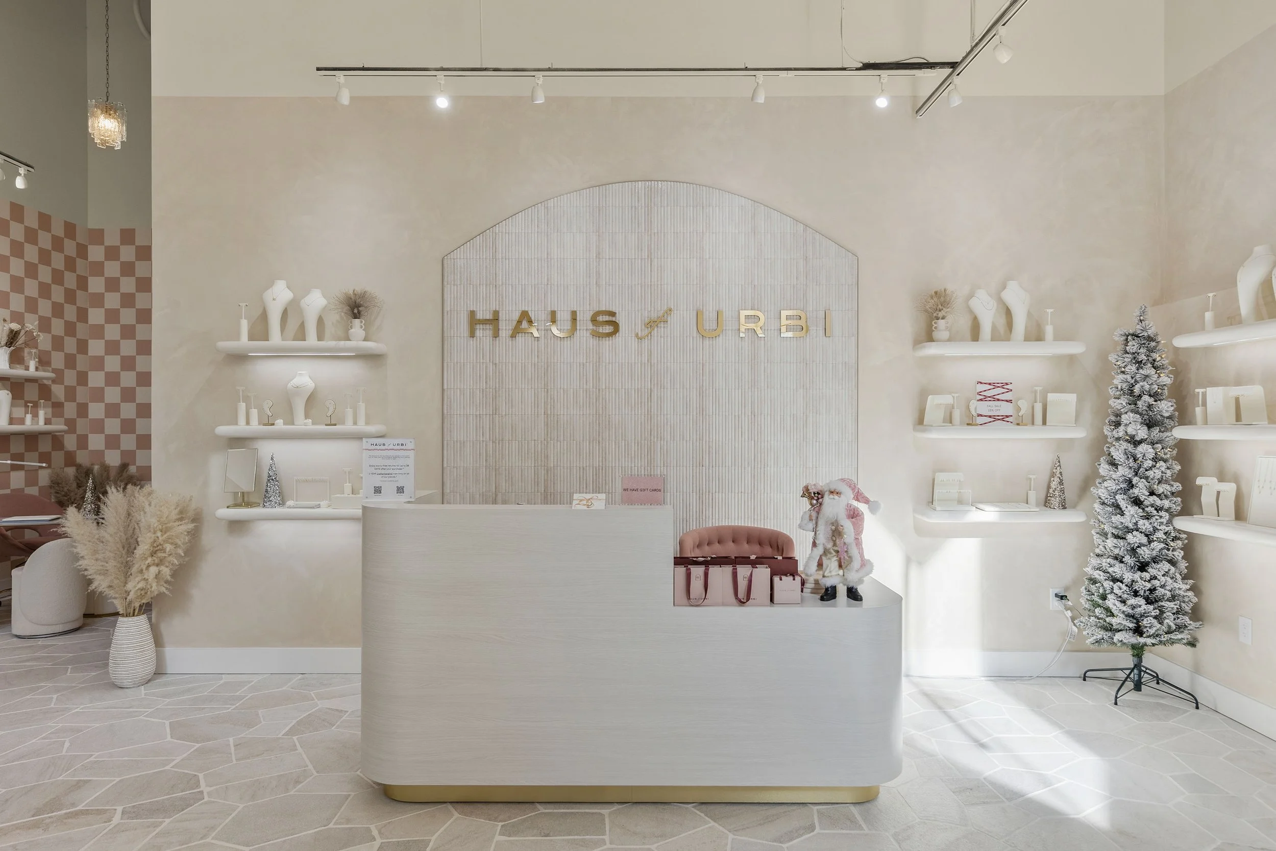 Interior of a retail store with a modern, minimalistic design. There is a curved white reception desk with pink and beige handbags on top. Behind the desk is a wall with the text 'HAUS of URBI' in gold letters. The store features decorative white she
