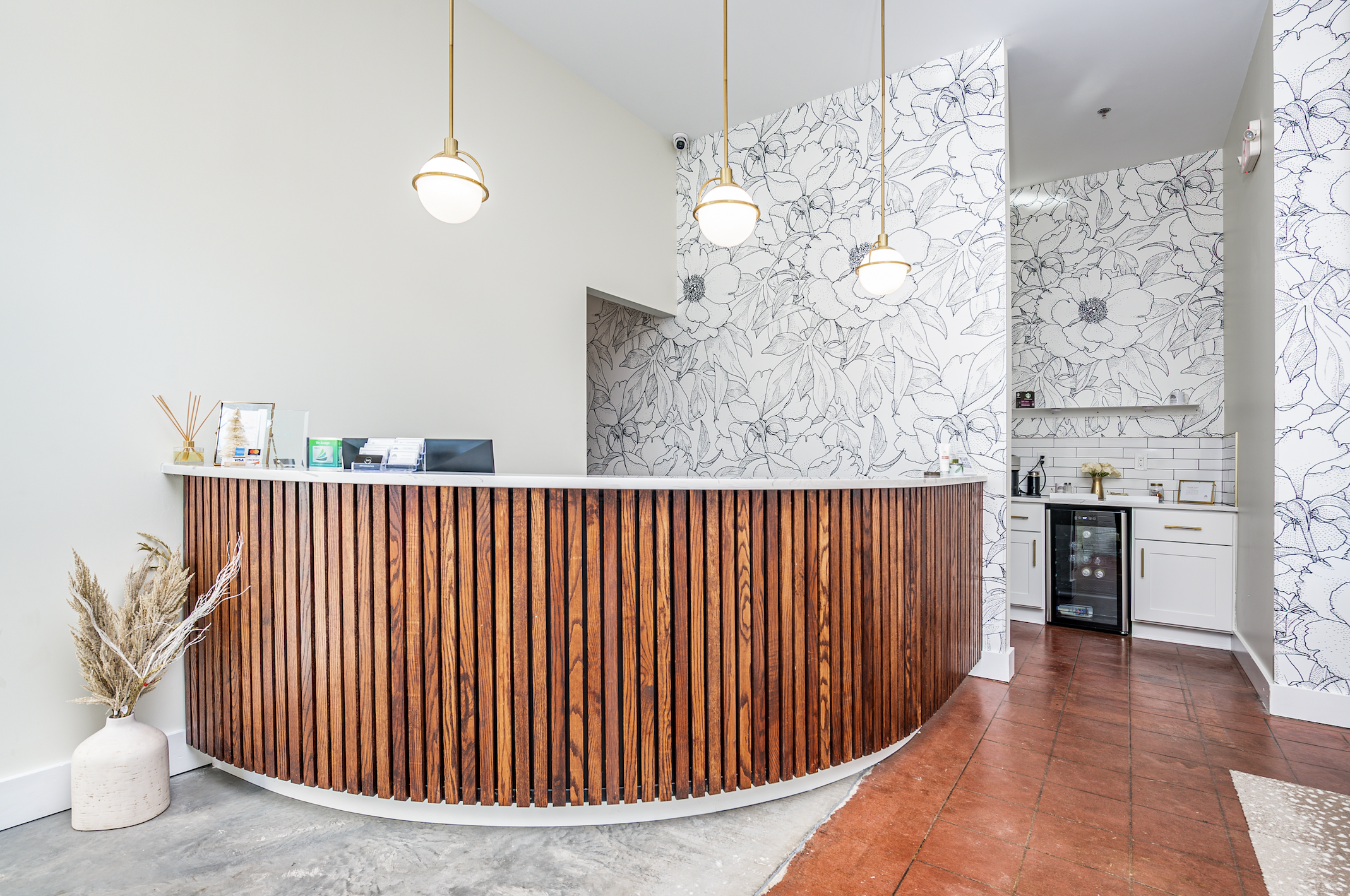 Modern reception area with a curved wooden counter, hanging pendant lights, white wall with floral wallpaper, small wine fridge, and minimal decor.