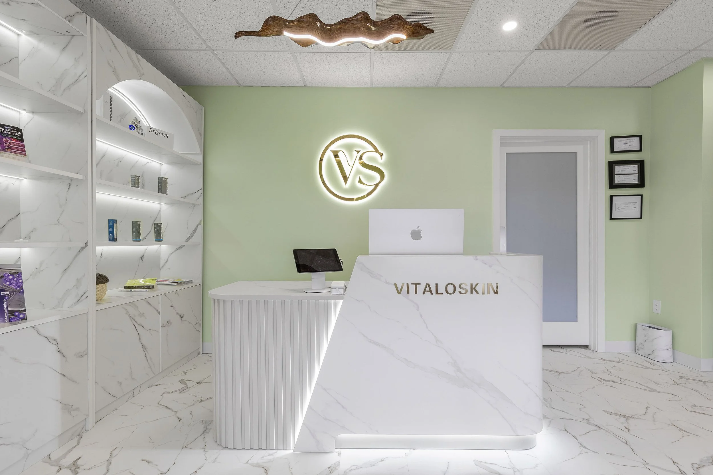 Reception area of VitaloSkin clinic with a white marble front desk, an Apple iMac computer, and a wall sign with 'VS' logo illuminated. Shelves with skincare products and framed certificates are visible, with light green walls and marble flooring.