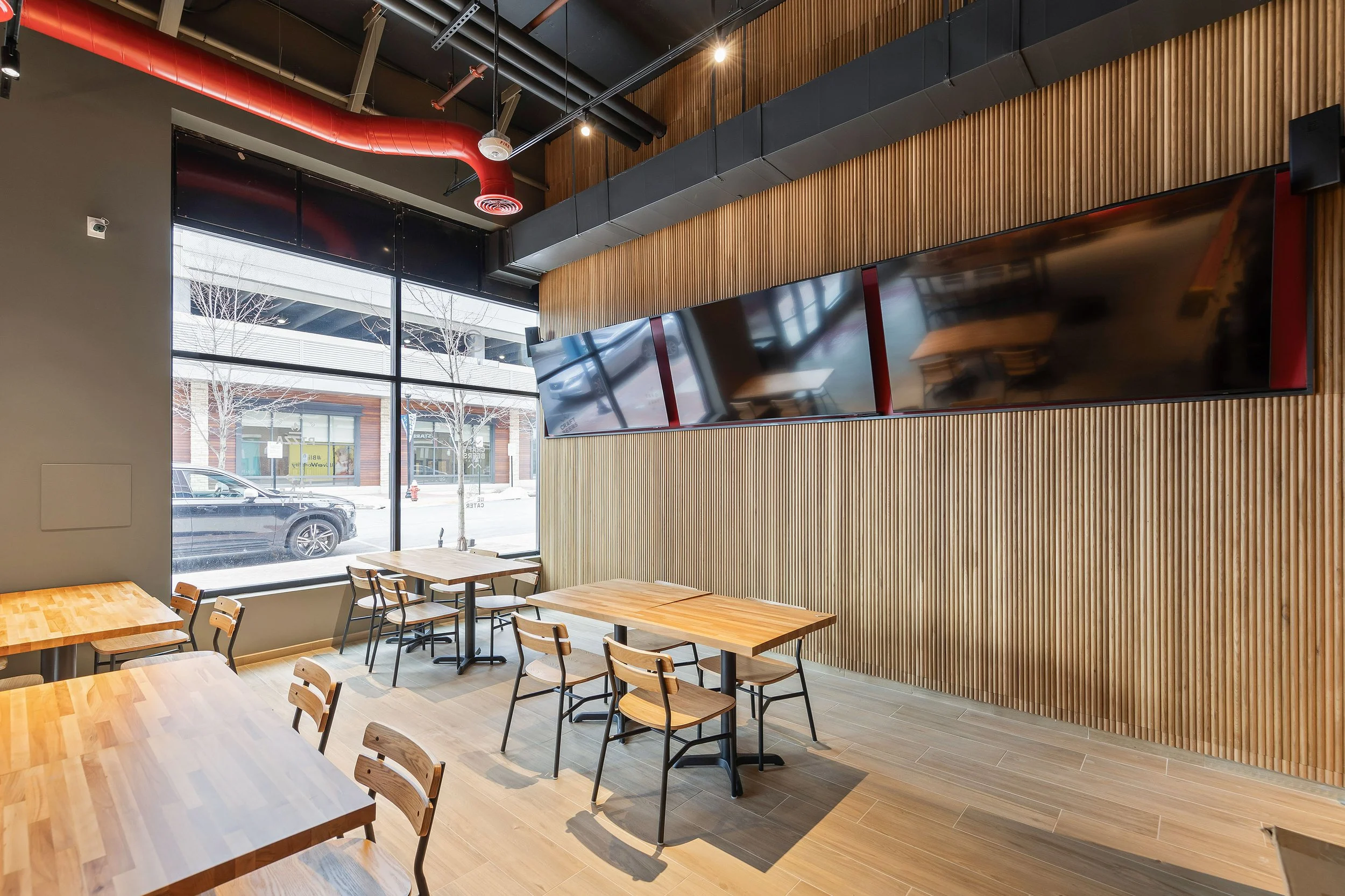 Interior of a modern restaurant or cafe with wooden tables and chairs, large window showing street view, three large mounted TVs on a wooden-paneled wall, exposed ductwork and ceiling pipes in industrial style.