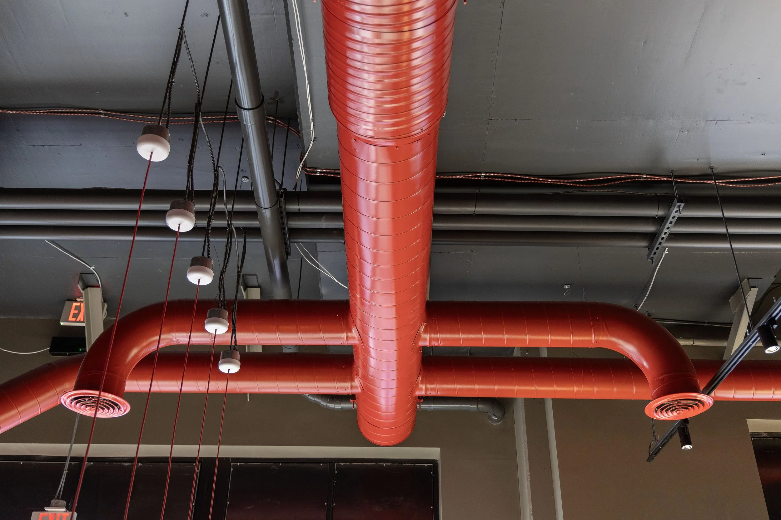Red industrial air ducts and pipes running along a gray ceiling, with some black conduit and hanging lights in an indoor space.