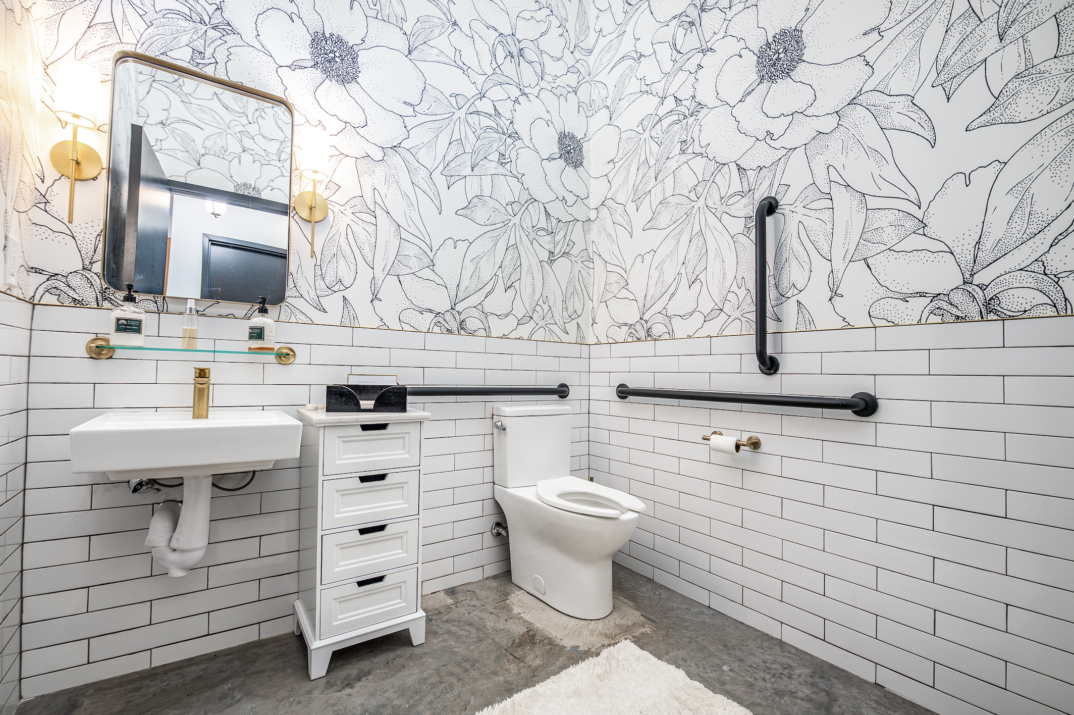 A modern bathroom with white subway tiles on the walls and black grout. The upper part of the wall features black and white floral wallpaper. There is a small white sink with a gold faucet, a mirror above the sink, and a white storage unit with drawe