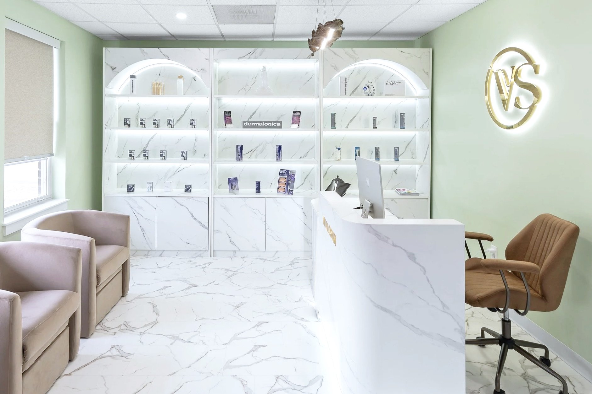 Interior of a modern clinic or office with white marble flooring and walls. There are beige and brown chairs near a white reception desk with a computer. Behind the desk, white marble shelves display skincare or cosmetic products with illuminated lighting, and a sign reading 'dermalogica'. On the right wall, there is a illuminated gold logo with the letters 'VS'.
