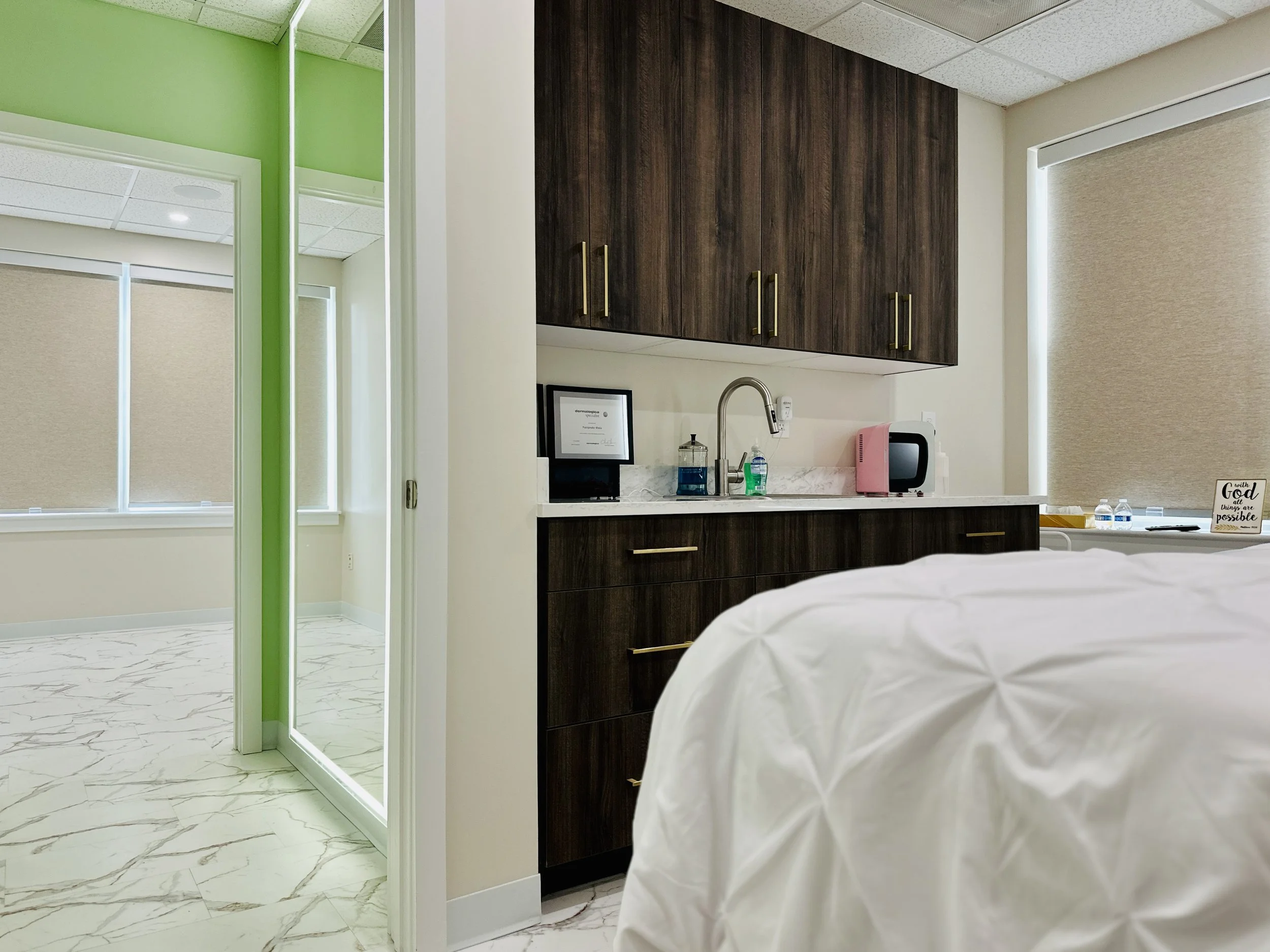 Hospital room with a bed covered in white sheets, a kitchenette with dark wood cabinets, a sink, small appliances including a microwave and water bottles, and windows with beige blinds.