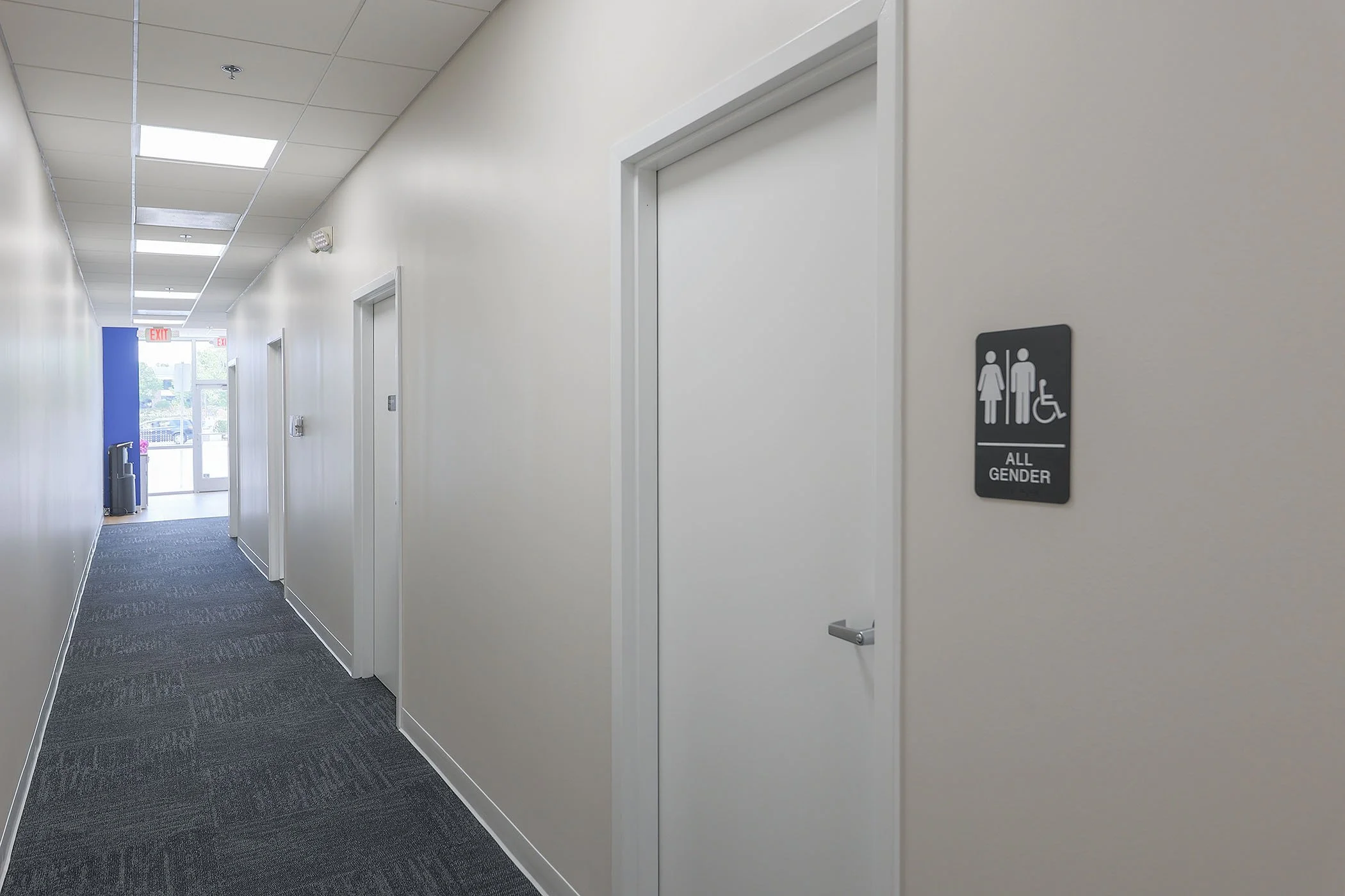 Hallway in a public building with beige walls, gray carpet, and a sign indicating an all-gender restroom.