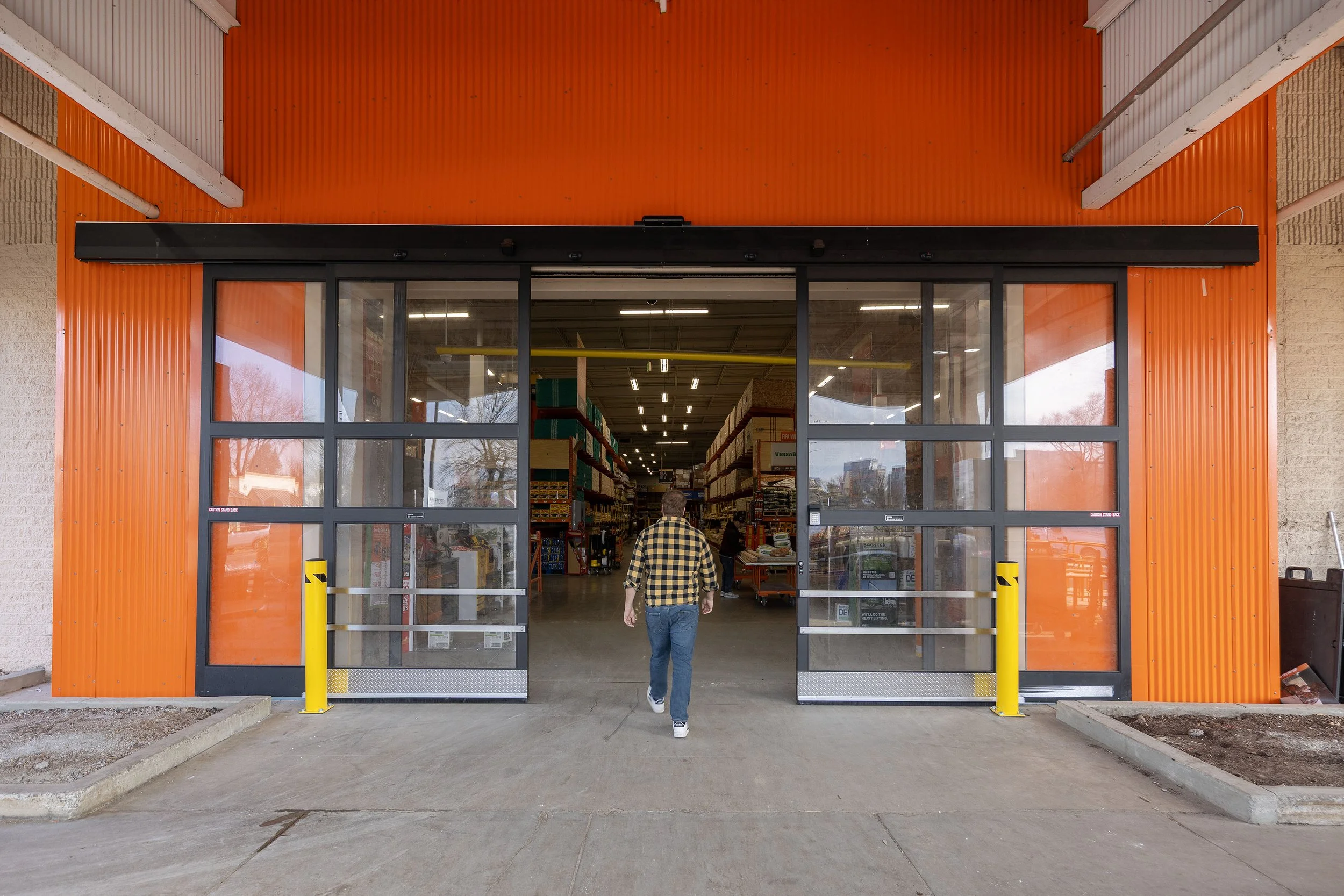 A person entering a hardware store with open glass doors, orange exterior walls, and shelves stocked with various products inside.